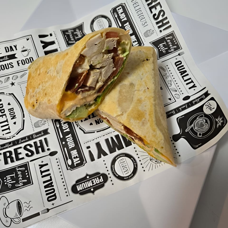 A chicken and bacon wrap, cut in half, rests on deli-themed parchment paper.