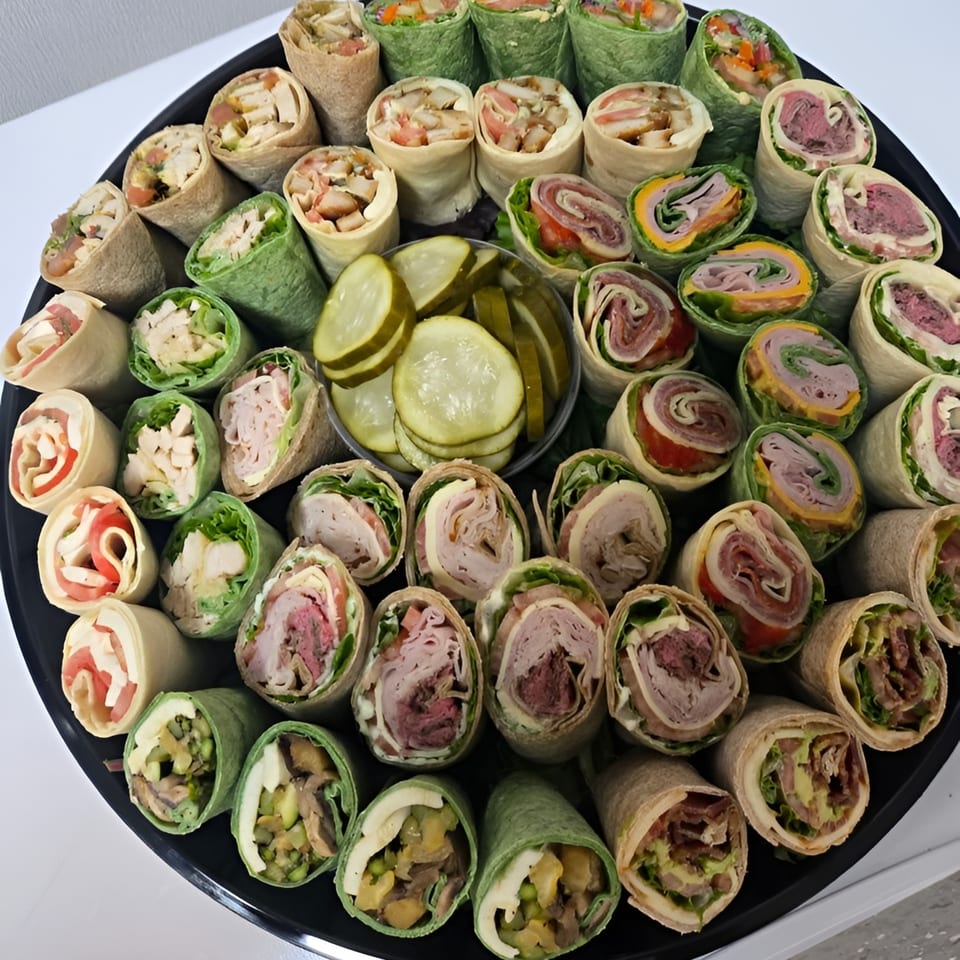 A catering tray of assorted deli wraps, including meat, cheese, and vegetable fillings, with pickles.